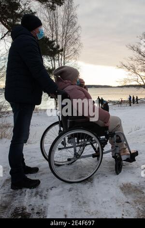Un uomo si prende cura di una donna su una sedia a rotelle. Sono fuori per una passeggiata. Serata invernale. Indossano maschere mediche. Foto di alta qualità Foto Stock