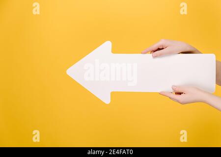 Primo piano vista laterale di belle mani femminili tenendo grande freccia di carta bianca, isolato su sfondo giallo studio con spazio di copia per la pubblicità Foto Stock
