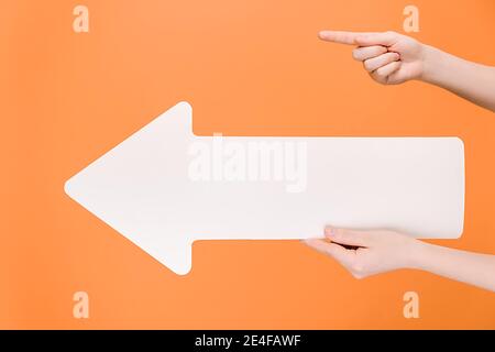 Immagini orizzontali di mani femminili che tengono una grande freccia di carta bianca, con il dito che punta allo spazio di copia per la pubblicità, isolate su sfondo arancione studio Foto Stock