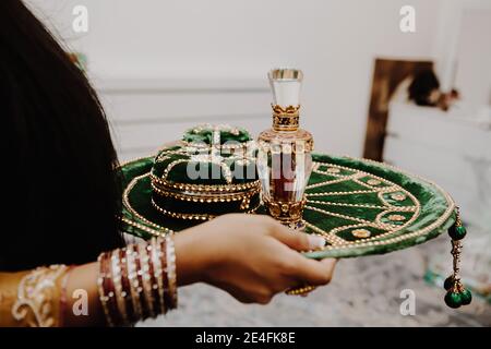 Mani femminili con vassoio indiano rotondo di velluto verde smeraldo dorato - piatto di nozze mehndi con henné in scatola e profumo primo piano del flacone Foto Stock