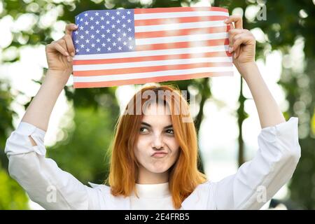 Ritratto di divertente giovane donna con capelli rossi che tiene la bandiera nazionale degli Stati Uniti in piedi in piedi all'aperto nel parco estivo. Ragazza positiva che festeggia United sta Foto Stock