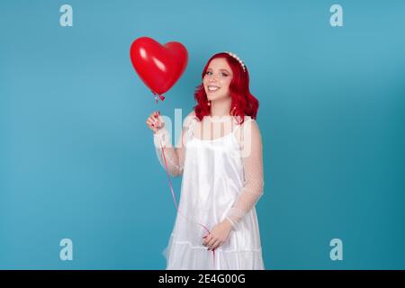 una giovane donna ridente e allegra in un vestito bianco con capelli rossi tiene in mano un pallone volante a forma di cuore, isolato su uno sfondo blu Foto Stock