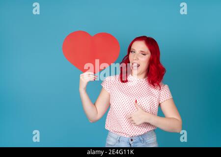 felice, gioiosa giovane donna con i capelli rossi tiene il cuore di carta rossa, gli verricelli, la lingua di messa fuori e dà i pollici su isolato su sfondo blu Foto Stock