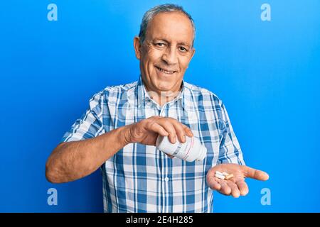 Uomo ispanico anziano che tiene pillole sorridenti con un sorriso felice e fresco sul viso. Mostrando i denti. Foto Stock