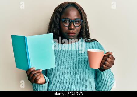 Giovane donna africana che legge un libro e beve una tazza di caffè depressa e si preoccupa di angoscia, piangendo arrabbiato e impaurito. Espressione triste. Foto Stock