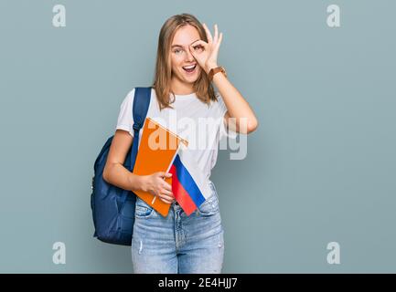 Bella giovane donna bionda scambio studente tenendo la bandiera russa sorridente buon fare ok segno con mano su occhio guardando attraverso dita Foto Stock