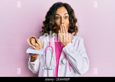 Media età medico ispanico donna che tiene modello anatomico di utero femminile con feto che copre la bocca con la mano, scioccato e impaurito per errore Foto Stock