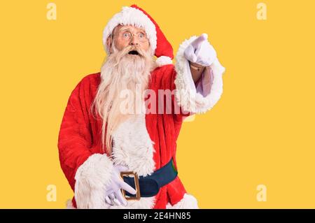 Vecchio uomo anziano con capelli grigi e barba lunga che indossa il costume tradizionale di babbo natale che punta con il dito sorpreso davanti, bocca aperta expressio stupito Foto Stock