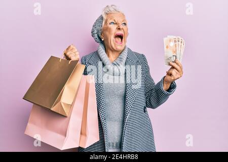 Donna anziana con capelli grigi che tiene borse per la spesa e banconote inglesi in sterline arrabbiate e pazza urlando frustrato e furioso, gridando con rabbia. Rabbia e un Foto Stock