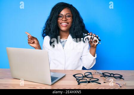 Bella donna africana che tiene occhiali di optometria sorridente felice indicando con mano e dito di lato Foto Stock