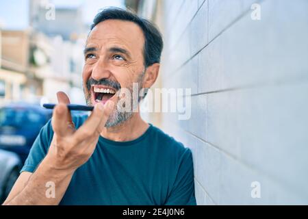 Uomo di mezza età con barba sorridente all'aperto allegro che invia voce sul telefono Foto Stock