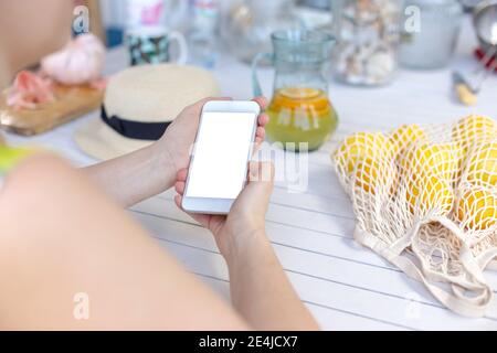 Mani di una ragazza che tiene uno smartphone bianco, su un tavolo di legno bianco, accanto ai limoni gialli in una borsa. Vista dall'alto. Foto Stock