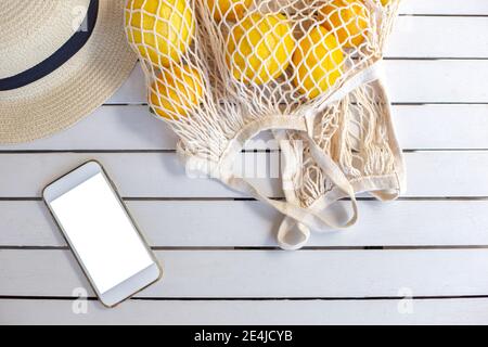 Uno smartphone bianco giace su un tavolo di legno bianco, accanto ad esso è una borsa a maglia con limoni e un cappello di paglia. Vista dall'alto Foto Stock