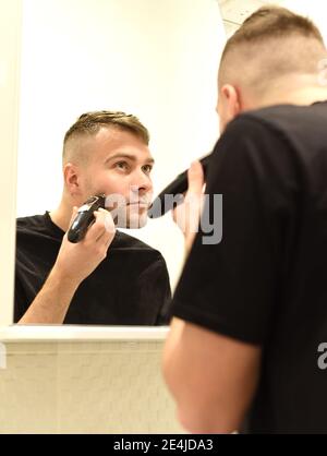 Rasatura di un giovane uomo con rifinitore e sguardo nello specchio in bagno Foto Stock