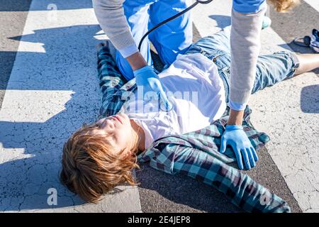 Operaio di salvataggio maschile con stetoscopio che esamina ragazzo sdraiato su strada dopo un incidente stradale Foto Stock