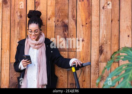 Bella donna millennial utilizzando smartphone da scooter elettrico push contro muro di legno Foto Stock