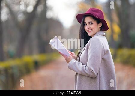 Sorridente bella donna che legge libro mentre si è in piedi nel parco durante fine settimana Foto Stock