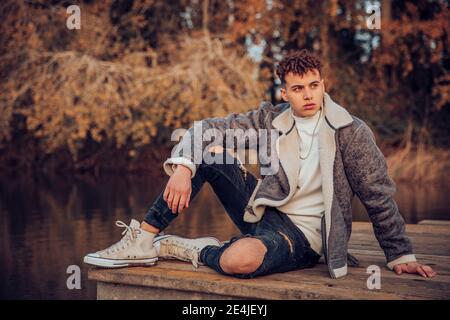 Giovane uomo che indossa una giacca seduto sul molo contro il lago a. autunno foresta Foto Stock