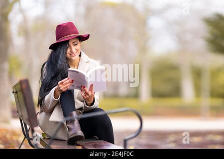 Bella donna con lunghi capelli libro di lettura mentre si siede sopra panca nel parco Foto Stock