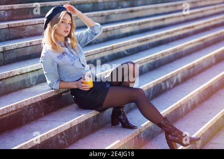 Donna bionda con beretto, bevendo un succo d'arancia naturale in un bicchiere di cristallo, seduto su alcuni gradini. Foto Stock