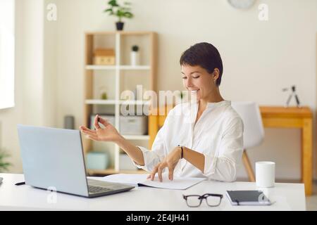Allenatore sorridente che ha una lezione online con uno studente o un webinar di hosting seduto alla scrivania con computer portatile Foto Stock
