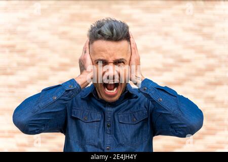 Uomo frustrato coprendo le orecchie mentre gridando contro il muro Foto Stock