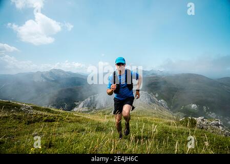 Percorso sportivo maturo che corre in montagna sul prato contro il cielo blu Foto Stock