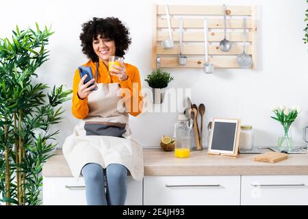 Donna sorridente che usa il telefono cellulare mentre beve succo d'arancia seduto sul tavolo a casa Foto Stock