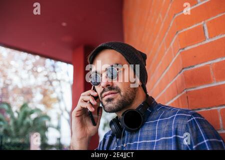 Giovane uomo alla moda che indossa occhiali da sole sulla chiamata telefonica contro il mattone parete Foto Stock