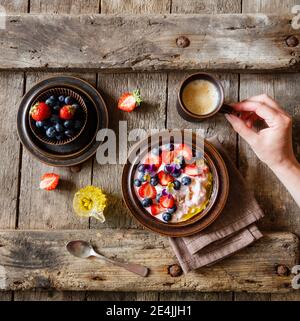 Mano femminile che raggiunge la tazza di caffè accanto alla piastra di quark con frutta e fiori commestibili Foto Stock