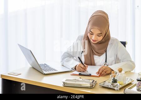 Donna musulmana sicura medico medico seduto alla scrivania, scrivendo su blocco note con computer portatile in clinica ospedale ufficio Foto Stock