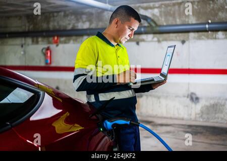 Tecnico maschile che utilizza il telefono cellulare con il computer portatile in auto elettrica in officina di riparazione auto Foto Stock