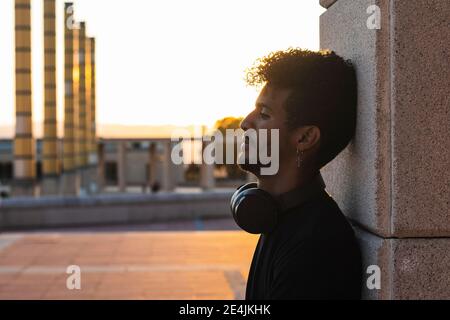 Giovane uomo attento con le cuffie appoggiate alla parete contro la luce cielo durante il tramonto Foto Stock