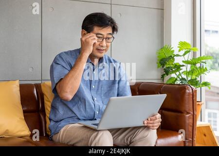 Felice asiatico anziano pensionato uomo seduto sul divano, utilizzando un computer portatile Foto Stock