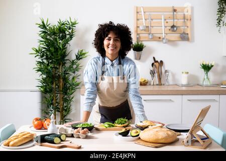 Ritratto di giovane donna appoggiata sul tavolo da cucina e sorridente alla telecamera Foto Stock