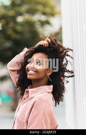 Donna afro sorridente con la mano in capelli mentre si sta in piedi città Foto Stock