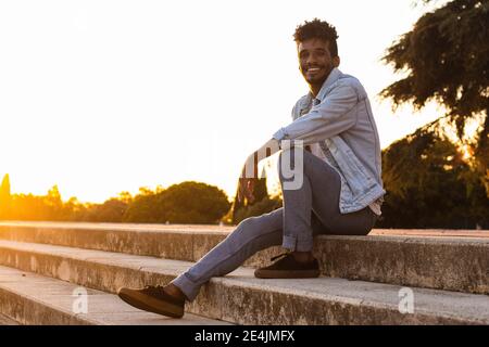 Ragazzo sorridente che indossa una giacca in denim seduta su gradini cielo limpido al tramonto Foto Stock