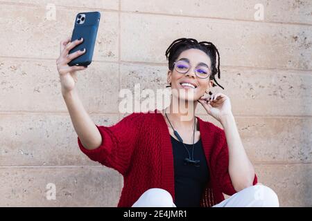 Felice bella giovane donna con Dreadlock che prende selfie contro muro Foto Stock