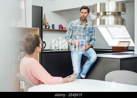 Uomo maturo che tiene la tazza di caffè mentre parla con la donna dentro cucina a casa Foto Stock