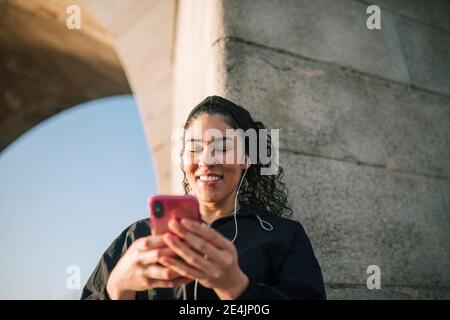 Donna sportiva sorridente che usa il telefono cellulare mentre ascolta la musica dalla parete Foto Stock