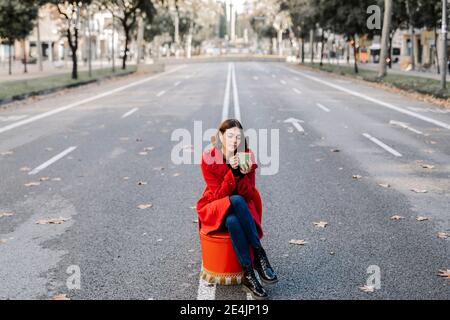 Giovane donna alla moda che tiene una tazza di caffè mentre ascolta la musica con occhi chiusi sulla strada Foto Stock