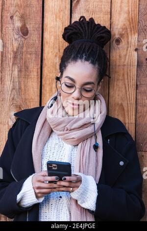 Bella giovane donna che usa lo smartphone mentre si appoggia contro il legno parete Foto Stock