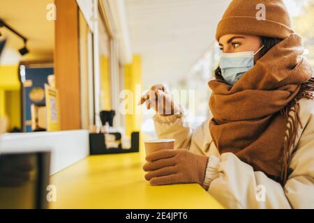 Giovane donna che indossa una maschera con una tazza di caffè usa e getta in strada cafe durante l'autunno Foto Stock