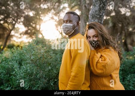 Giovane donna che indossa la maschera facciale abbracciando l'uomo da dietro mentre In piedi alla foresta durante Covid-19 Foto Stock