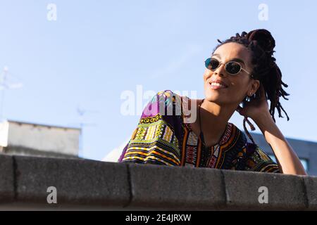 Donna con dreadlock che indossa occhiali da sole che guardano lontano mentre si appoggia parete di ritegno contro il cielo Foto Stock
