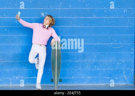 Giovane alla moda con skateboard che prende selfie mentre si alza in piedi gamba contro parete blu Foto Stock