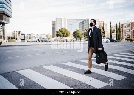 Uomo d'affari che indossa giacca e maschera che tiene la valigia mentre cammini su strada in città Foto Stock