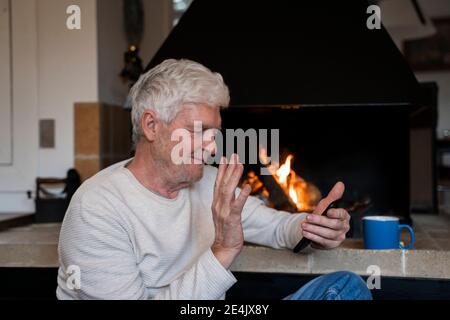 Uomo anziano che sventolava la mano per videochiamare sul telefono cellulare mentre si siede a casa Foto Stock