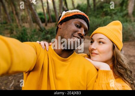 Uomo che indossa un cappello a maglia che prende selfie con la donna mentre si è in piedi alla foresta Foto Stock