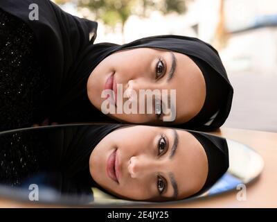 Giovane bella donna che indossa hijab nero riflettendo in specchio rotondo Foto Stock